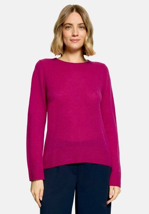 Femme aux cheveux blonds mi-longs portant un pull rose vif à manches longues et un pantalon foncé sur un fond uni.