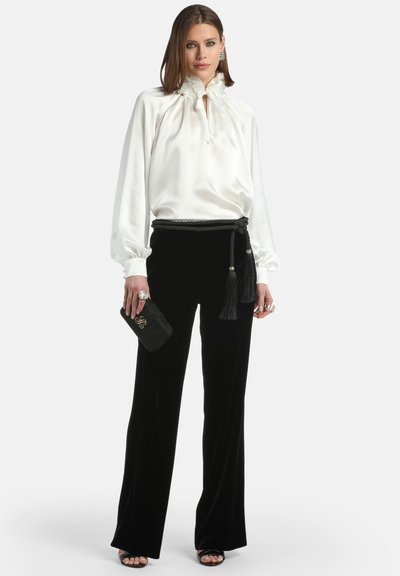 Blusa di raso bianca con colletto alto arricciato, abbinata a pantaloni di velluto neri con cintura intrecciata e dettaglio con frange. Accessorio a borsa a mano nera.