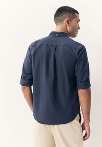 Navyblauw overhemd met knopen en opgerolde mouwen, voorzien van een rugpas en subtiele stikdetails. Gemaakt van een lichtgewicht stof.