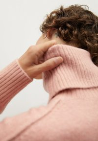 Personne aux cheveux bouclés portant un pull côtelé rose remonte son col haut, révélant un petit tatouage noir derrière l'oreille sur un fond uni.