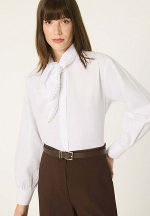 Femme aux cheveux bruns et raides portant un chemisier blanc avec un col décoratif noué et un pantalon marron taille haute avec une ceinture.