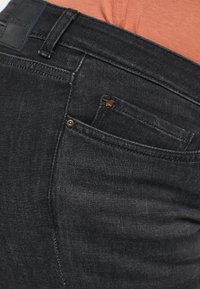 Svarta denimjeans har framträdande sömmar, en liten logotyp på fickan och en slät textur. Midjebandet har en läderlapp.