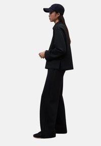 Mujer con un atuendo completamente negro, pantalones de pierna ancha, blusa de manga larga y gorra negra, de perfil sobre un fondo blanco.