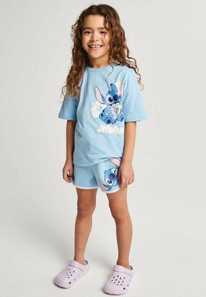 Lächendes Kind mit langen lockigen Haaren, das ein hellblaues T-Shirt und Shorts mit Stitch-Motiv trägt und barfuß vor einem einfarbigen Hintergrund steht.
