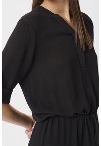 Blusa nera leggera con scollo a V, maniche corte arrotolate e vita elastica. Il tessuto ha una consistenza morbida e increspata.