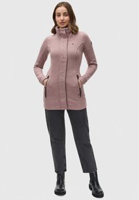 Veste rose à manches longues avec col haut, poches zippées et coupe ajustée, assortie à un jean foncé et des bottines noires.