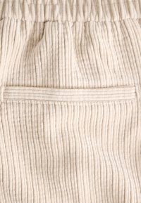 Béžová kordová tkanina s vertikálnymi ryhami, so viditeľnou vreckovou časťou integrovanou do dizajnu. Mäkká textúra a elastický pás.