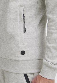 Lys grå hettegenser med en sidelomme med svart glidelås, teksturert stoff, ribbede mansjetter og en sirkulær svart logo detalj på nederste kant.