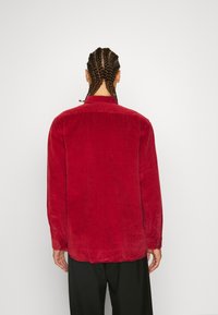 Camisa de pana roja con yugo posterior, mangas largas, puños de botón y dobladillo recto. Tejido texturizado con estrías verticales.