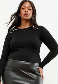 Zwarte longsleeve trui met decoratieve knopen op de schouder en een getailleerd ontwerp, gecombineerd met een zwarte faux-lederen rok.