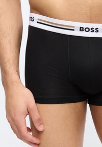 Torse masculin et haut des cuisses portant un boxer noir avec une ceinture blanche portant l'inscription « BOSS », main reposant près de la hanche.