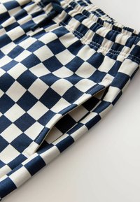 Tuvaļskats uz tumši zilu un baltu rūtainu elastīgu jostasvietas biksēm ar redzamu sānu kabatu un šuvju detaļām.