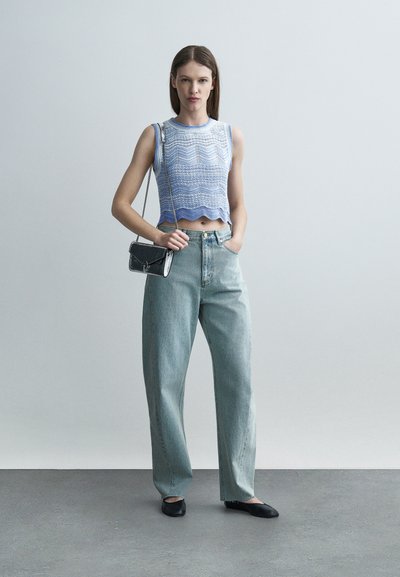 Γυναίκα που φοράει μπλε πλεκτό αμάνικο τοπ, τζιν με ψηλή μέση και φαρδιά μπατζάκια, μαύρα μπαλαρίνες, και κρατά μια μικρή μαύρη τσάντα στον ώμο, μπροστά σε απλό φόντο.