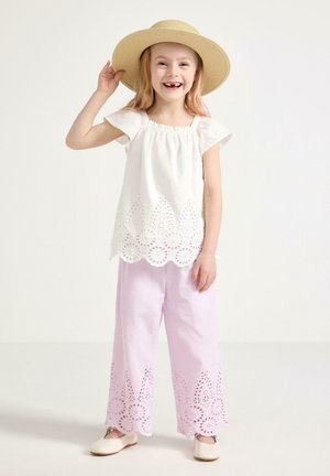 Smilende ung pige iført en bredskygget stråhat, hvid bluse med hulmønster, lyserøde bukser med hulmønster og cremefarvede ballet flats mod en ensfarvet baggrund.