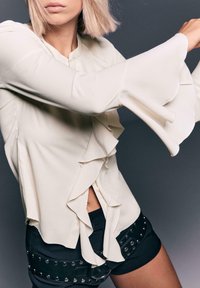 Femme portant un chemisier en crème avec des volants et des manches évasées, ainsi qu'une ceinture cloutée noire sur un short noir, posant devant un fond gris foncé.