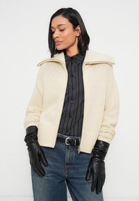 Μπεζ πλεκτή ζακέτα με ριμπ λεπτομέρειες και ψηλό γιακά, συνδυασμένη με μαύρο ριγέ πουκάμισο και μαύρα δερμάτινα γάντια.