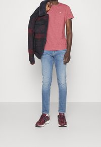 Červené heather tričko s kulatým výstřihem v kombinaci s modrými džíny, tmavě kostkovanou flanelovou bundou a bordó teniskami s bílými akcenty.