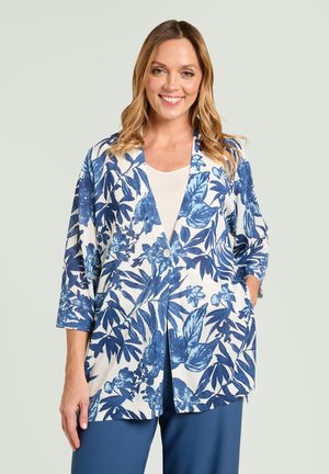 Lächelnde Frau mit welligem blondem Haar, die einen weißen Blazer mit blauem Blumenmuster, ein weißes Oberteil und blaue Hosen trägt, Hände in den Taschen.