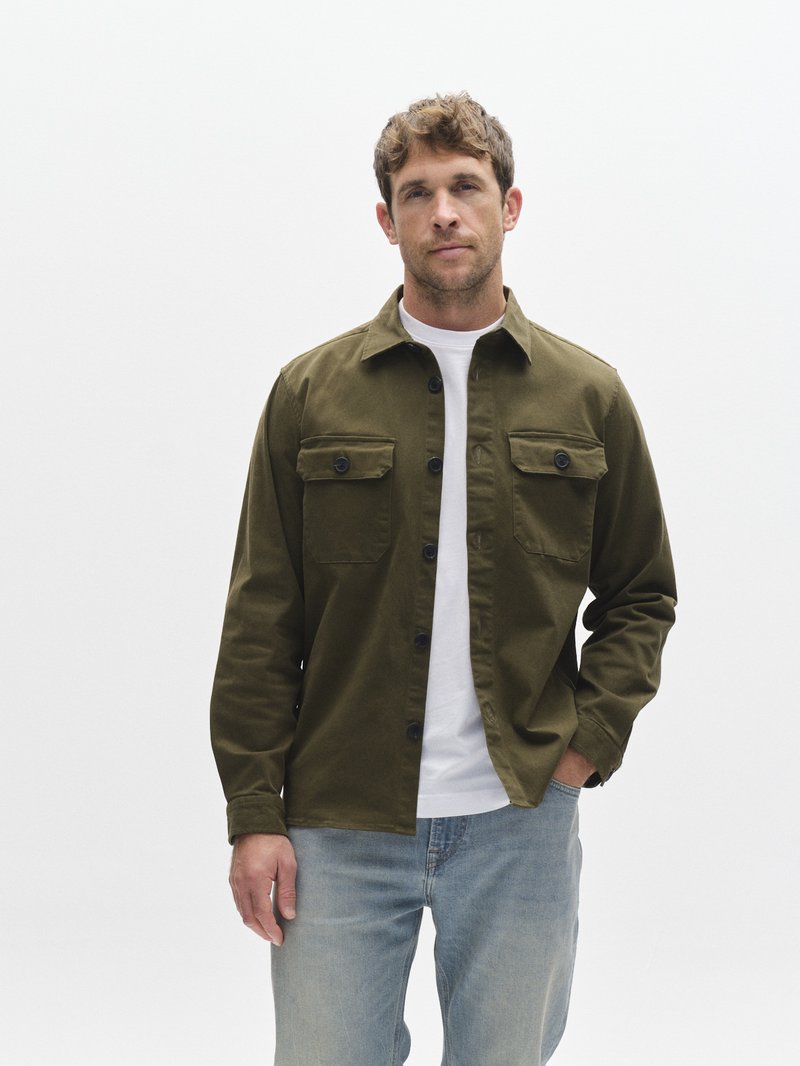 Olivovo zelená košeľa na zapínanie s dvoma náprsnými vreckami, čierne gombíky a jednoduché biele tričko pod ňou, v kombinácii so svetlomodrými džínsami.
