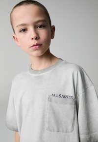 Světle šedé tričko s krátkým rukávem, které má na přední straně kapsu a nad ní v tmavém písmu nápis "ALLSAINTS". Materiál z jemné bavlny.