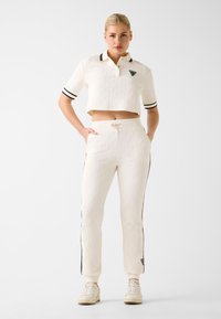 Biała krótka koszulka polo z fakturowanym wzorem, czarnymi paskami i trójkątnym logo; do tego białe spodnie z troczkiem i bocznymi paskami.