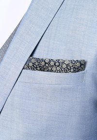 Blazer azzurro chiaro con un tessuto testurizzato. Il fazzoletto da taschino presenta un motivo floreale scuro con accenti in argento, parzialmente esposto nella tasca.