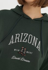 Donkergroene hoodie met capuchon, voorzien van geborduurde tekst "ARIZONA", "WILD WEST" en een ontwerp van cowboylaarzen. Zacht materiaal met een trekkoord in de capuchon.