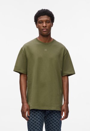Camiseta verde oliva de gran tamaño hecha de algodón texturizado. Presenta un cuello redondo y un sutil detalle del logo en el pecho. Combinada con pantalones oscuros estampados.