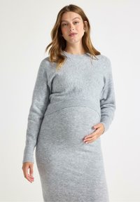 Gravid kvinna klädd i en mysig grå tröja och klänning, som varsamt håller om sin mage, med en neutral bakgrund som framhäver hennes kläder.