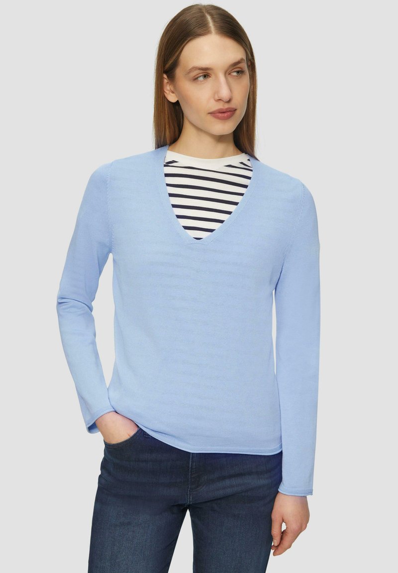 Helles blauer Pullover mit langen Ärmeln und V-Ausschnitt, aus weichem Material. Über einem gestreiften Hemd getragen, kombiniert mit dunklen Jeans.