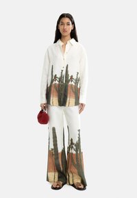 Komplet z lnu składający się z koszuli i spodni z nadrukiem krajobrazu pustynnego z kaktusami, w odcieniach kremowych i ziemnych. Dodatkiem jest mała czerwona torba.