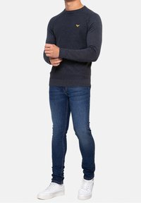 Hombre vestido con un suéter de manga larga gris oscuro con un logo amarillo, jeans ajustados azules y zapatillas blancas, de pie con los brazos cruzados.