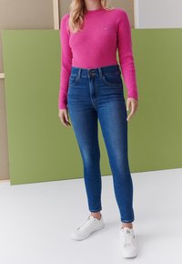 Top de mangas largas acanalado en fucsia combinado con jeans skinny azules de cintura alta y zapatillas blancas, con un fondo verde.