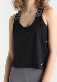 Černý sportovní top s výstřihem do V a šedými značkovými ramínky. Vyrobený z hladkého materiálu, má zkrácený střih a bílé logo na přední straně.