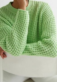 Personne portant un pull tricoté épais vert clair, reposant son bras et sa main sur une surface blanche, fond neutre.