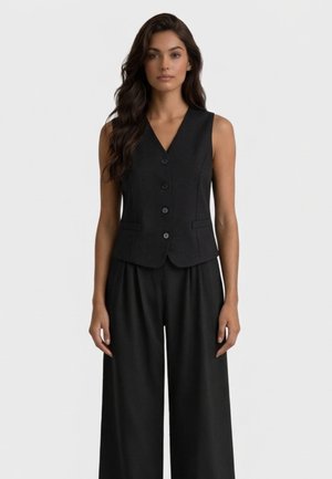 Donna con lunghi capelli scuri che indossa un gilet nero senza maniche con bottoni e pantaloni neri a gamba larga, in piedi davanti a uno sfondo chiaro uniforme.