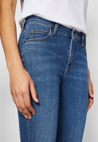 Jeans de mezclilla azul con un corte ajustado, que presentan bolsillos frontales y una cinturilla. La tela tiene una textura suave con detalles de decoloración sutil.