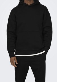 Sudadera con capucha negra, con un bolsillo tipo canguro, mangas anchas y ribetes acanalados. Presenta un pequeño logo en el pecho y una textura suave.