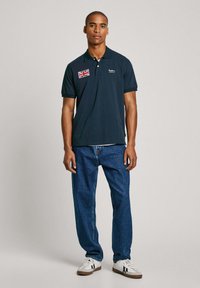Marineblauw poloshirt met korte mouwen, een kraag, Britse vlag patch, gecombineerd met losse blauwe jeans en witte sneakers.