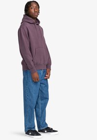Felpa viola con cappuccio e coulisse, abbinata a jeans blu a taglio morbido e sneakers nere con dettagli a stella bianca.