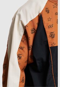 Farvet jakke med orange, hvid og sorte paneler. Den orange sektion har et mønster af sorte logoer og stjerner. Glat tekstur.