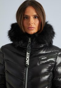Melnā pūkainā jaka ar spīdīgu tekstūru, aprīkota ar noņemamu mākslīgās kažokādas kapuci, ar rāvējslēdzēju un izteiksmīgu "JACKIT" zīmolu priekšpusē.