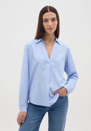 Giovane donna con capelli lisci e scuri indossa una camicetta azzurro chiaro a maniche lunghe e jeans blu, in piedi con una mano in tasca.