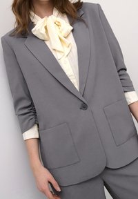 Blazer gris au design structuré, avec des revers à encoches et deux poches avant. Porté sur un chemisier crème avec un nœud ruban au col.