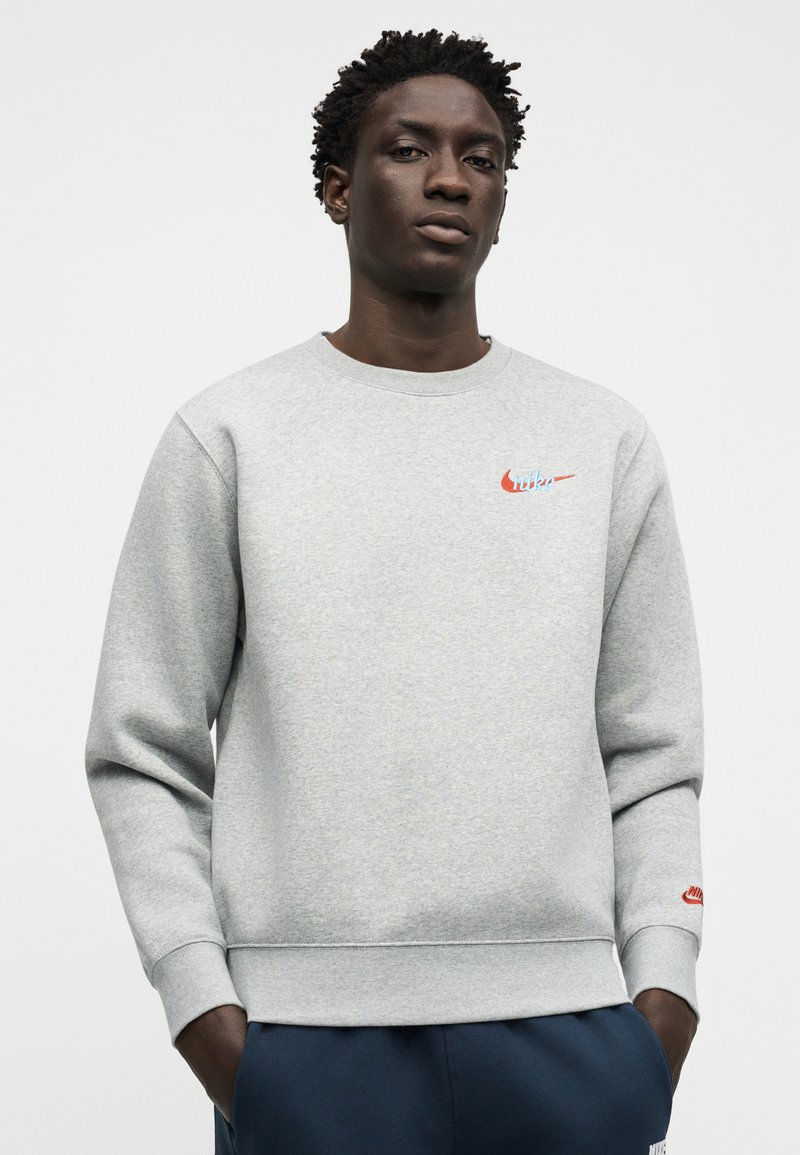 Young Black man wearing a light gray Nike sweatshirt with red logos, standing with hands in navy blue pants pockets against a plain background.