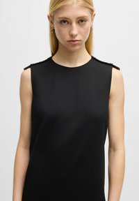 Schwarzes ärmelloses Kleid mit hohem Kragen, aus glattem Stoff gefertigt. Es hat kleine Schulterakzente und eine figurbetonte Silhouette.