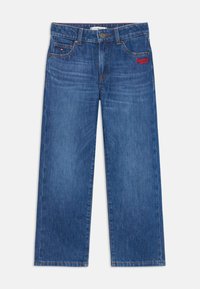 Vaqueros de denim azul con un diseño de pierna recta, que presentan costuras visibles y un pequeño logo rojo en el lado izquierdo. Textura levemente desgastada.