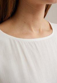 Vit, lätt blus med en slät textur och rund halsringning, försedd med diskreta vertikala randningar för extra detalj.