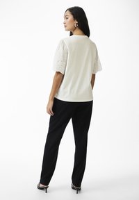 Top alb cu mâneci scurte, umflate, cu dantelă; designul din spate este simplu. Acesta este asortat cu pantaloni negri, largi și încălțăminte cu toc.