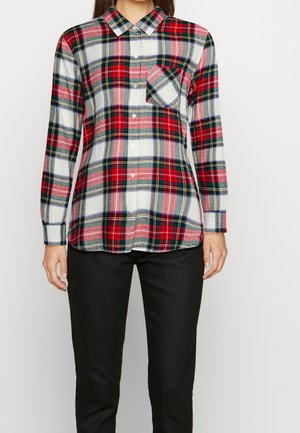 Woman wearing a red, white, and black plaid button-up shirt with long sleeves and black pants, standing against a plain background.